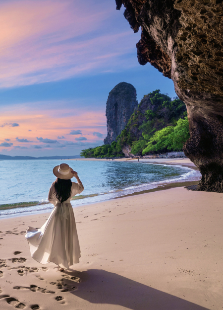 A woman in a white dress stands at the entrance of a cave on Railay Beach, Krabi, Thailand, with the ocean in the background.